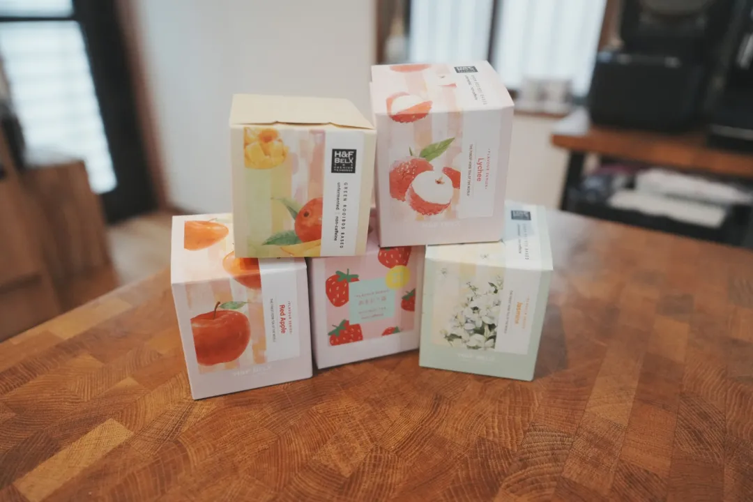 H&F BELX 루이보스티 상자 다섯 개를 나무 테이블 위에 쌓아 둔 모습, 리치·레드 애플·재스민·아마오우 딸기·망고 그림